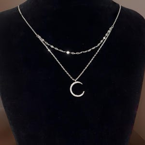 Könnte beinhalten: Eine silberne Halskette mit mehreren Ebenen und einem Mondsichel-Anhänger. Die Halskette hat zwei zarte Ketten, wobei die untere Kette einen Mondsichel-Anhänger mit kleinen, funkelnden Steinen trägt. Der Hintergrund ist schwarz.