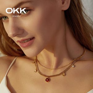 Puede incluir: Un collar dorado de dos capas, con un colgante rojo y pequeños abalorios. El collar lo lleva una persona de piel clara. La marca "OKK Jewelry" es visible en la esquina superior izquierda.