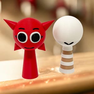 Puede incluir: Dos peque&ntilde;as figuritas impresas en 3D. Una es roja con cuernos y ojos grandes, la otra es blanca con una base a rayas marrones y blancas.