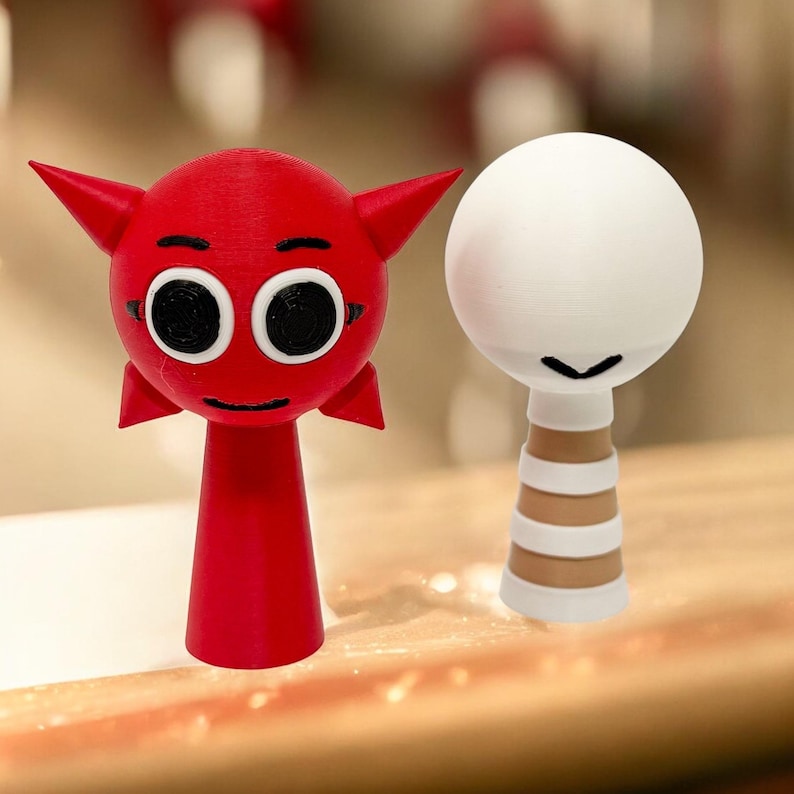 Puede incluir: Dos peque&ntilde;as figuritas impresas en 3D. Una es roja con cuernos y ojos grandes, la otra es blanca con una base a rayas marrones y blancas.