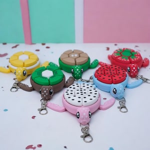 May include: Six colorful turtle-shaped keychains with different designs. Each keychain has a removable top that reveals a different pattern, including a kiwi, a strawberry, a polka dot pattern, a cookie, and a heart pattern.
