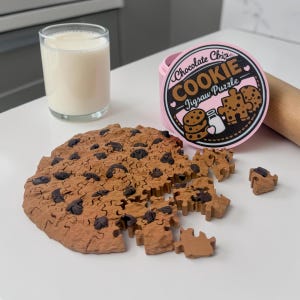 May include: A chocolate chip cookie jigsaw puzzle with a glass of milk. The puzzle is brown with dark chocolate chip details. The puzzle pieces are scattered around the cookie. A pink container with the words "Chocolate Chip Cookie Jigsaw Puzzle" is also in the image.