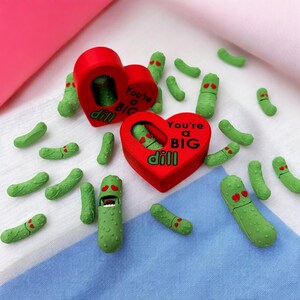May include: Two red heart-shaped boxes with the text "You're a BIG dill" inside. The boxes are surrounded by green pickle-shaped figurines with faces.