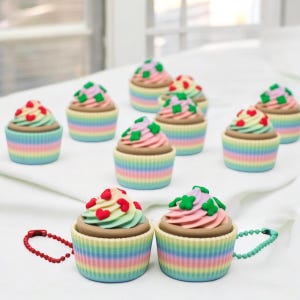 Op de afbeelding: Een set kleurrijke cupcake-sleutelhangers met regenboog-glazuur en verschillende toppings, waaronder harten en klavertjesvier. De sleutelhangers zijn gemaakt van plastic en hebben een metalen ketting.