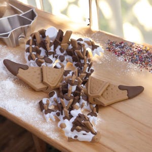 May include: A whimsical, multi-coloured cookie cutter set arranged on a wooden surface. The set includes a metal cookie cutter and a decorative cookie shaped like a dinosaur with brown, white, and tan frosting and chocolate pieces.