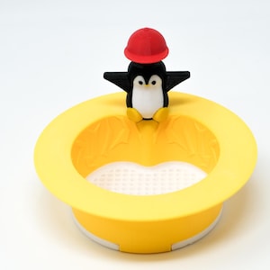 May include: A yellow silicone sink strainer with a penguin figurine wearing a red hat on top. The strainer has a white mesh bottom.