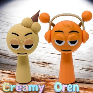Puede incluir: Dos figuritas impresas en 3D con caras de dibujos animados. La figurita de la izquierda es amarilla con un cono de helado marr&oacute;n en la cabeza. La figurita de la derecha es naranja con una diadema y auriculares. El texto "Creamy Oren" est&aacute; impreso debajo de las figuritas.