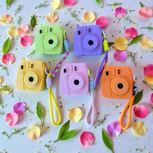 Puede incluir: Una colección de seis cámaras instantáneas de colores pastel, incluyendo verde, morado, amarillo, rosa y naranja. Cada cámara tiene una correa de muñeca a juego. El fondo está decorado con pétalos de flores y vegetación.