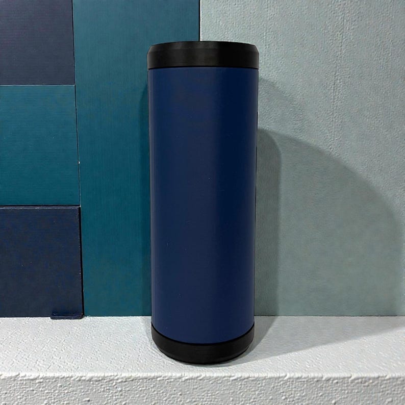 May include: A cylindrical beverage cooler featuring a matte navy blue body with a solid black rim at the top and a matching black base. The container has a smooth, uniform finish and a vertical orientation, designed to hold a standard beverage can.