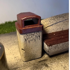 Puede incluir: Un pequeño cubo de basura de cemento con tapa granate y cuerpo gris claro. El cuerpo tiene texto estilo grafiti. El cubo de basura está sobre una superficie de hormigón.