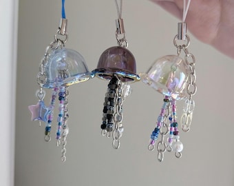 May include: Three handmade jellyfish charms with iridescent, purple, and blue bell-shaped tops. Each charm has a chain and beaded tentacles in coordinating colors, with a star or teardrop charm. They are attached to silver-toned metal loops.