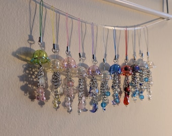 May include: A collection of colorful, decorative charms hanging from a white bar. Each charm features a clear, bell-shaped top with a dangling chain of beads and charms in various colors, including pink, blue, and green. The charms are suspended by colorful cords.
