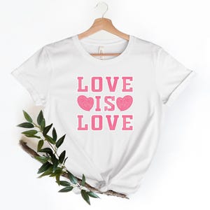 Può includere: Maglietta bianca con il testo "LOVE IS LOVE" in rosa con due cuori rosa.