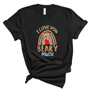 Può includere: Maglietta nera con una grafica arcobaleno e il testo "I love you beary much". La grafica presenta un orsacchiotto che tiene un cuore.