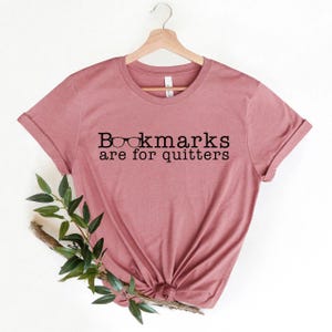 Può includere: Una maglietta rosa polverosa con il testo "Bookmarks are for quitters" e una grafica di occhiali.