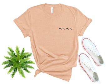 以下が含まれることがあります： 前面に黒い筆記体で「mama」と書かれたピーチ色のTシャツです。