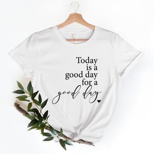 Può includere: Maglietta bianca con testo nero che recita "Today is a good day for a good day" con un piccolo cuore alla fine.