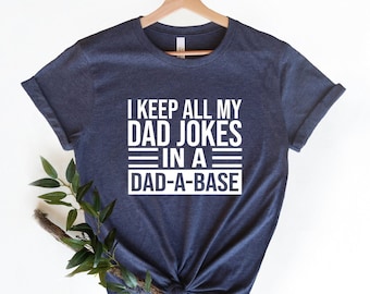 May include: A dark heather navy blue short-sleeve t-shirt featuring white text that reads I KEEP ALL MY DAD JOKES IN A DAD-A-BASE with horizontal lines separating the phrases. The shirt is displayed on a wooden hanger.