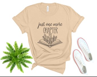 May include: A beige t-shirt with the text "just one more chapter" and a black and white illustration of a book with flowers on top.
