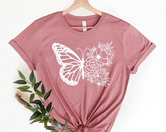 May include: A mauve short-sleeve t-shirt featuring a white graphic print on the front. The design consists of a butterfly on the left side and a cluster of various flowers on the right side, creating a symmetrical composition.