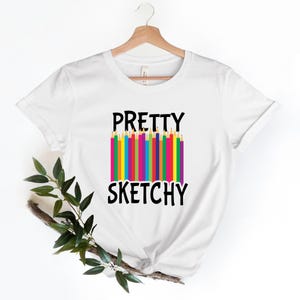 Può includere: T-shirt bianca con la scritta "PRETTY SKETCHY" in nero. Tra le parole c'è una fila di matite colorate. La maglietta è appesa a una gruccia di legno.