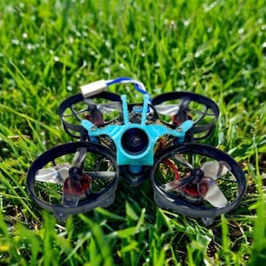 May include: A small, quadcopter drone with a turquoise frame and black propellers rests on a bed of green grass. The drone has four propellers and a camera. A white and blue cable extends from the top.