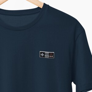 May include: A navy blue t-shirt with a small, embroidered retro video game controller. The controller design includes a directional pad, buttons, and a rectangular shape. The shirt is made of a soft material.