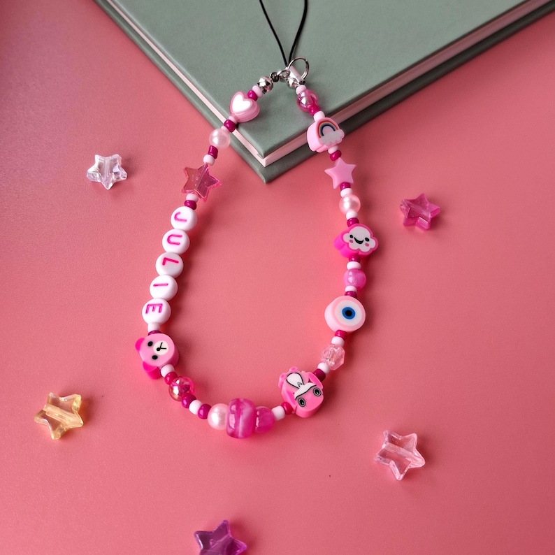 Personalised pink phone charm with pink letter beads. Y2K aesthetic style pink name charm lying on a pink background and on the corner of a book. Perfect gift for teen girls.