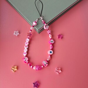 Personalised pink phone charm with pink letter beads. Y2K aesthetic style pink name charm lying on a pink background and on the corner of a book. Perfect gift for teen girls.