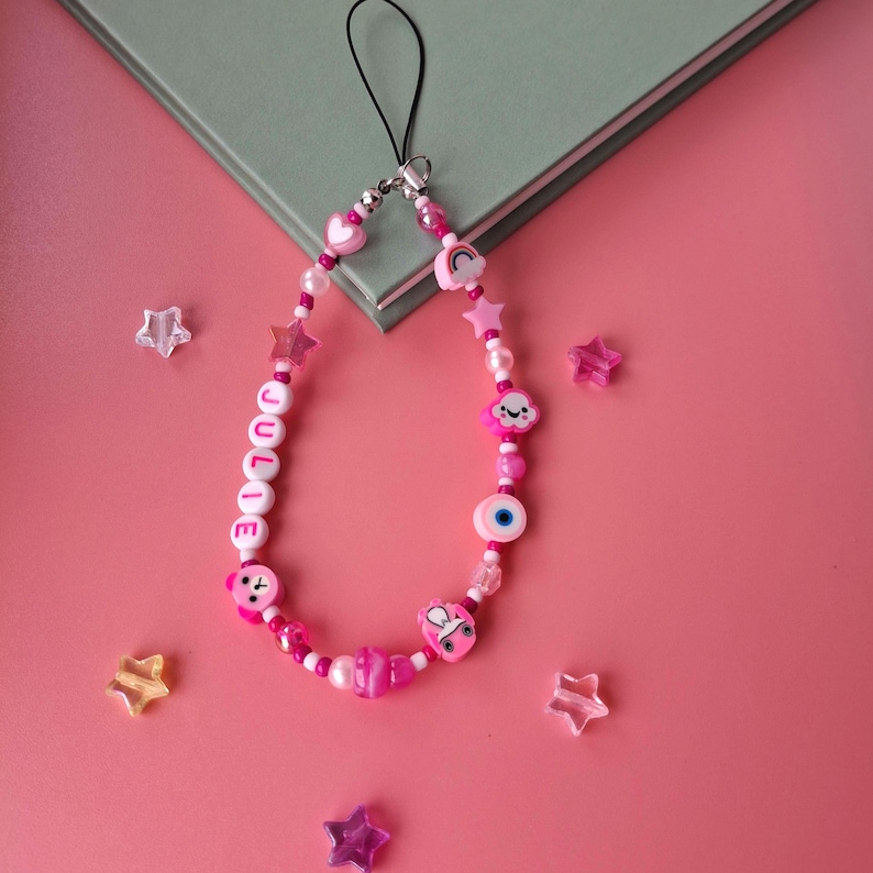 Personalised pink phone charm with pink letter beads. Y2K aesthetic style pink name charm lying on a pink background and on the corner of a book. Perfect gift for teen girls.