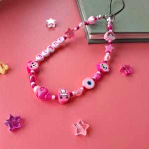 Personalised pink phone charm with pink letter beads. Y2K aesthetic style pink name charm lying on a pink background and on the corner of a book. Perfect gift for teen girls.