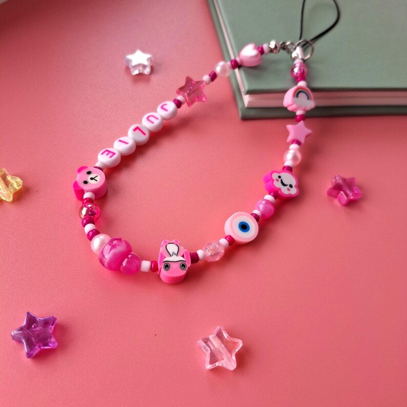 Personalised pink phone charm with pink letter beads. Y2K aesthetic style pink name charm lying on a pink background and on the corner of a book. Perfect gift for teen girls.