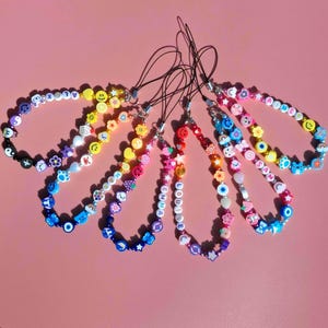 以下が含まれることがあります： カラフルなビーズの携帯電話チャームのコレクション。各チャームには、スマイリーフェイス、花、ハート、星など、さまざまなビーズが使用されています。 「LOUISE」や「SAM」などの名前が書かれたチャームもあります。チャームは黒いコードに付いています。