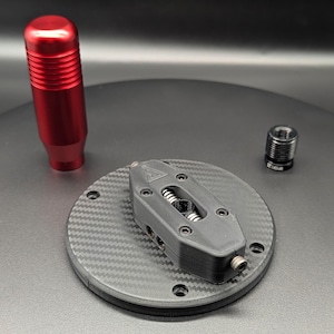 Könnte beinhalten: Ein roter zylindrischer Schaltknauf mit horizontalen Rillen, eine schwarze kreisförmige Montageplatte mit Carbonfaser-Muster und zentralem mechanischem Bauteil sowie ein kleiner schwarzer Gewindeadapter aus Metall für den Einbau.