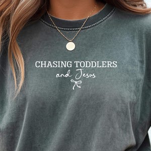 May include: Dark gray t-shirt with the text "CHASING TODDLERS and Jesus" in white. The shirt has a relaxed fit and a crew neck. The text is centered on the front of the shirt.