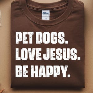 May include: A brown t-shirt with the words "PET DOGS. LOVE JESUS. BE HAPPY." in white block letters. The shirt is folded neatly and is surrounded by neutral-toned objects, including a cup of coffee and twine.