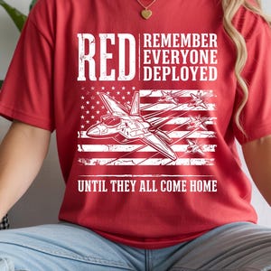 May include: Red t-shirt with white text and a graphic of a fighter jet over an American flag. The text reads "RED REMEMBER EVERYONE DEPLOYED UNTIL THEY ALL COME HOME."