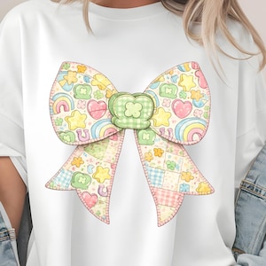 Puede incluir: Camiseta blanca con un gran diseño de lazo colorido. El lazo está decorado con arcoíris, corazones, estrellas y tréboles. El lazo tiene un patrón de patchwork con colores pastel. La camiseta es de estilo casual y oversize.