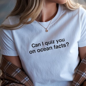 May include: White t-shirt with the text "Can I quiz you on ocean facts?" in black. The shirt is worn with a brown and tan plaid shirt. The model is wearing a gold necklace.