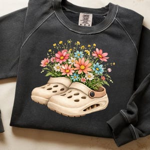 Op de afbeelding: Een zwarte sweatshirt met een afbeelding van beige klompen gevuld met kleurrijke bloemen. De bloemen zijn onder meer roze, blauwe, gele en witte madeliefjes. De sweatshirt heeft een ronde hals en lange mouwen.