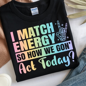 May include: Black t-shirt with the text "I MATCH ENERGY SO HOW WE GON' Act Today?" in a rainbow gradient. The design includes a skeleton hand making a rock and roll gesture. The shirt is displayed with sneakers and jeans.