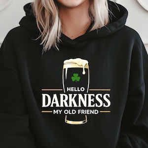 May include: Black hoodie with the text "HELLO DARKNESS MY OLD FRIEND" in white, with a beer glass graphic. The beer glass has a green shamrock and foam. The hoodie is worn by a person.