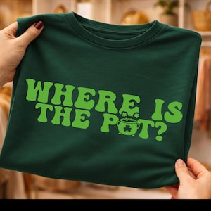 May include: A dark green t-shirt with the text "WHERE IS THE POT?" in bright green, featuring a pot of gold and a shamrock. The shirt is held up by hands, showcasing the design.