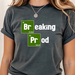May include: Dark gray t-shirt with the words "Breaking Prod" in white, with green periodic table-style boxes. The shirt has rolled-up sleeves and is paired with denim shorts.