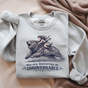 Puede incluir: Sudadera gris claro con un gráfico de una niña montada en un oso, con el texto azul marino "MAY OUR DAUGHTERS BE UNGOVERNABLE". La sudadera está doblada sobre una superficie rosa claro.