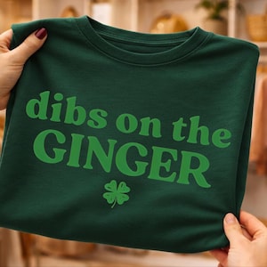 May include: A dark green crew neck t-shirt with the words "dibs on the GINGER" in green and a green four-leaf clover. The shirt is held up by hands.