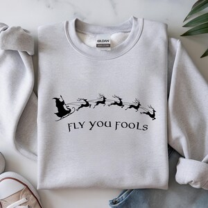 May include: A light gray sweatshirt with a black graphic of a sleigh pulled by reindeer, with the phrase "FLY YOU FOOLS" underneath. The sweatshirt has a crew neck and long sleeves.