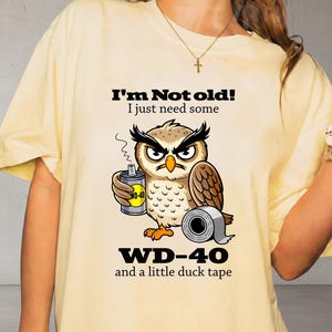 May include: Cream-colored t-shirt featuring a cartoon owl holding a WD-40 can and a roll of duct tape. The text on the shirt reads "I'm Not old! I just need some WD-40 and a little duck tape."