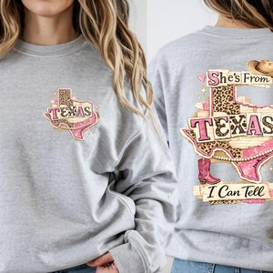 Może przedstawiać: Jasnoszara bluza z nadrukiem w kształcie Teksasu z napisem "TEXAS" w kolorze r&oacute;żowym i lamparcim. Tył bluzy zdobi większa grafika z napisem "She's From Texas I Can Tell", kapeluszem kowbojskim i butami.