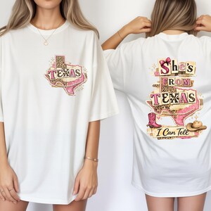 Może przedstawiać: Biała koszulka z grafiką stanu Teksas, butem kowbojskim i napisem "TEXAS" w kolorze r&oacute;żowym. Tył koszulki zawiera napis "She's From Texas I Can Tell" z butem kowbojskim i kapeluszem.
